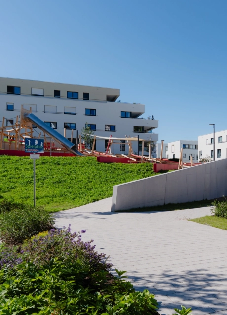 Spielplatz mit Rutschen und Klettergeräten aus Holz, umgeben von Grünflächen. Davor ist ein Weg aus grauem Pflastersteinen. Im Hintergrund sind moderne Wohnhäuser.