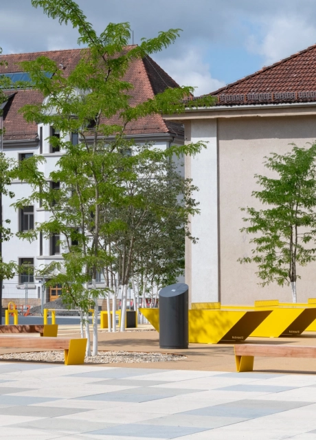 GDM.SCENIC individual auf einem großzügigen Platz mit gelben Sitzbänken, jungen Bäumen und offenen Aufenthaltsbereichen.