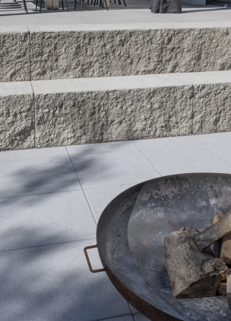 Granitfarbener GDM.MAXI block, welcher als zweistufige Sitzmöglichkeit dient. Auf Ihr sind verschiedene Kissen und in der Mitte ist eine Feuerschale mit Holz aus Metall. Auf der Bodenfläche ist GDM.NUEVA light verlegt.