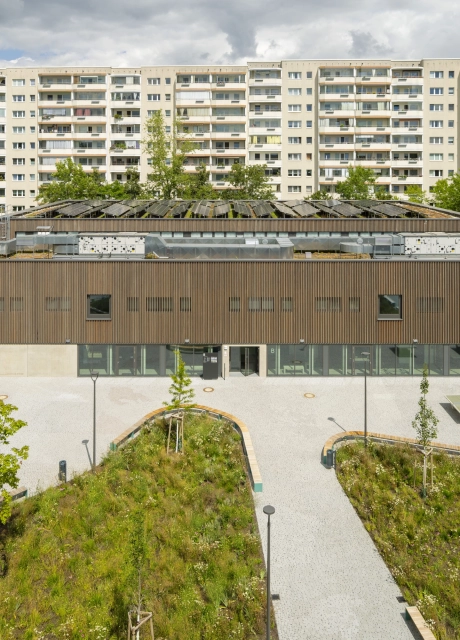 Luftaufnahme eines großen Schulgebäudes mit Holzfassade mitten in einer Stadt. Vor dem Gebäude liegt ein Schulhof mit vielen Sitzmöglichkeiten, Tischtennisplatten und begrünten Flächen.