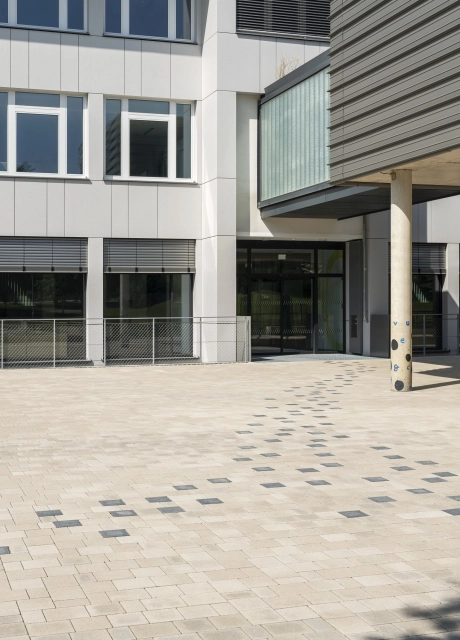 A path consisting of anthracite-colored concrete blocks leads through a beige paved schoolyard to a modern school building. The building has a partly white, partly light gray façade and a dark gray extension in the foreground. 