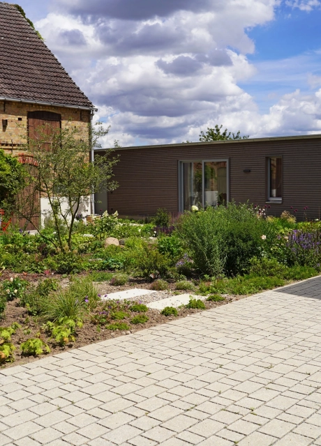 A rustic brick building with large wooden gates and a landscaped garden in the foreground. A paved path leads through the garden, which is planted with flowers and shrubs. In front of the garden is a natural beige paved courtyard laid out in rows on which a child is riding a bicycle. On the right is a modern building with large glass doors.