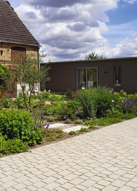 Ein rustikales Backsteingebäude mit großen Holztoren und einen angelegten Garten im Vordergrund. Ein gepflasterter Weg führt durch den Garten, der mit Blumen und Sträuchern bepflanzt ist. Vor dem Garten ist ein naturbeige gepflasterter Hof im Reihenverband angelegt auf welchem ein Kind auf dem Fahrrad fährt. Rechts ist ein modernes Gebäude mit großen Glastüren zu sehen.