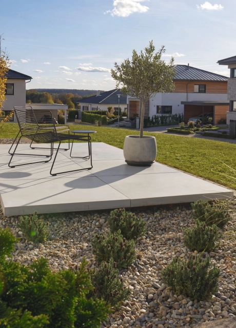 Moderne Terrasse mit hellen GDM.MASSIMO light Platten, die eine erhöhte Sitzfläche mit filigranen Metallstühlen bilden. Im Hintergrund fasst eine GDM.MASSIMO wall in Cortenstahloptik ein bepflanztes Hochbeet ein. Im Vordergrund liegen ein Kiesbeet mit Ziergräsern und immergrünen Pflanzen sowie eine angrenzende Rasenfläche in einem ruhigen Wohngebiet.