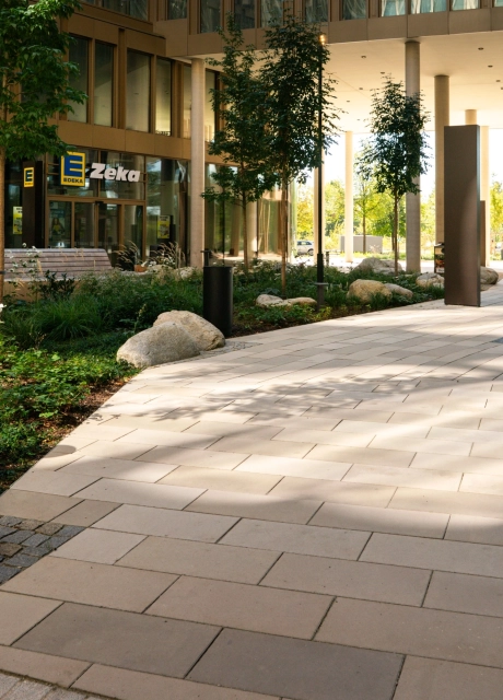Mit GDM.SCENIC individual gepflasterte Hoffläche in verschiedenen Kalksteinfarbtönen. In der Mitte befindet sich ein großes Beet mit Sträuchern, kleinen Bäumen und größeren Steinen. 
