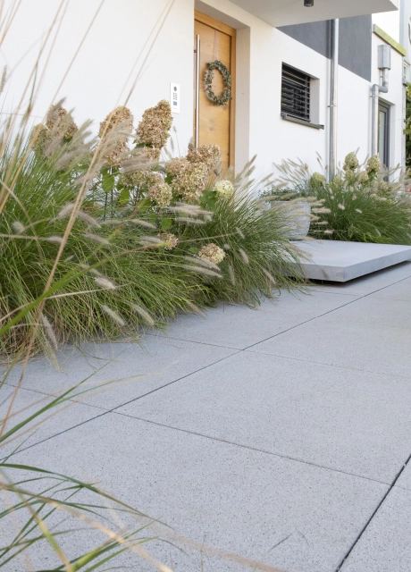 Graue Betonsteinplatten führen zum Hauseingang hin. Vor der Haustüre in Holzoptik ist ein graues Podest. Ringsherum sind Gräser und Sträucher.