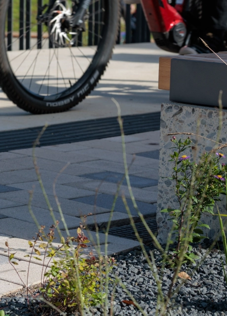 Nahansicht eines bepflanzten Bereichs mit Ziergräsern und dunklem Splitt im Vordergrund. Dahinter verläuft ein Weg aus beigem GDM.KLIMASTEIN, in den einzelne graue Pflastersteine als Akzente eingestreut sind. Rechts steht eine moderne Sitzbank mit einem Sockel aus Beton, im Hintergrund ist ein Fahrrad erkennbar.