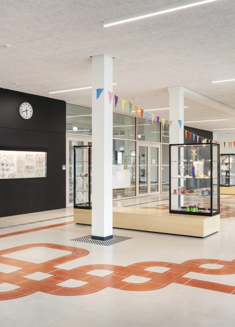 Der Eingangsbereich eines Schulgebäudes, durch dessen grau-beigen Schulboden sich ein rotes Band aus Sichtbeton-Platten zieht.