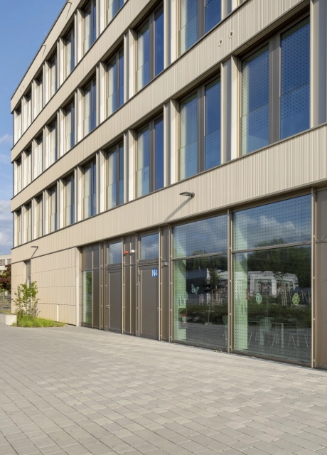 Fassade eines Schulgebäudes mit großen Fenstern. Davor ist ein grau-beigen geplasterter Platz und im Hintergrund eine begrünte Stadt mit vielen Bäumen zu erkennen.