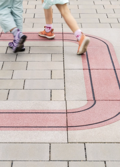 Nahaufnahme einer grau-beigen Pflasterfläche, durch welche sich ein Band aus roten Betonsteinen zieht. Die Beine zwei laufender Kinder sind drauf zu erkennen