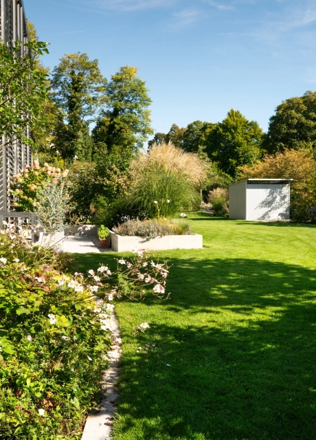 Gartenansicht mit großer Rasenfläche, dicht bepflanzten Beeten und einem Wohngebäude auf der linken Seite. In der Mitte steht ein kleines Beet mit Ziergräsern, während im Hintergrund ein Gartenhaus und zahlreiche Bäume unter blauem Himmel zu sehen sind.
