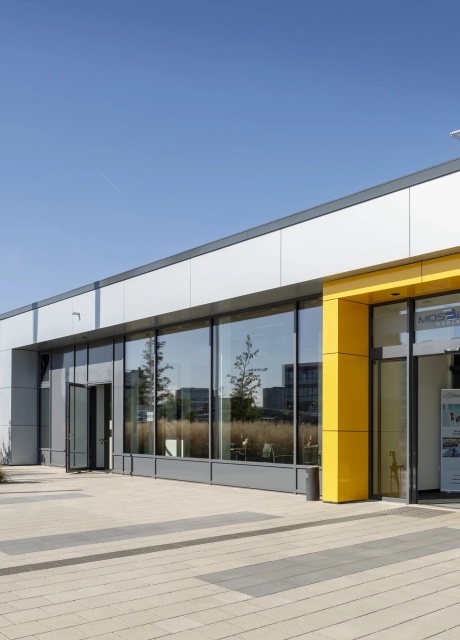 Gebäude mit grauer Fassade, großer Fensterfront und auffallender, gelber Umrahmung um die Eingangstür. Davor ist ein versickerungsfähiger, gepflasterter Platz aus grauem Betonstein. Daneben befindet sich helles Ziergras und zwei Fahnenmasten. 
