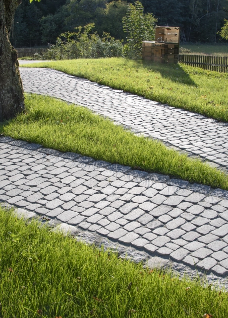 Sanft geschwungener Gartenweg aus sandfarbenem Betonstein, der durch eine gepflegte Rasenfläche zwischen Bäumen und einem Holzzaun verläuft.