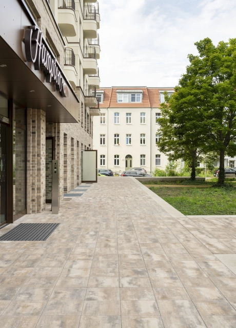 Gepflasterter, muschelkalkfarbener Gehweg entlang eines Gebäudes, daneben sind Grünflächen mit Bäumen und Wohnhäusern im Hintergrund.