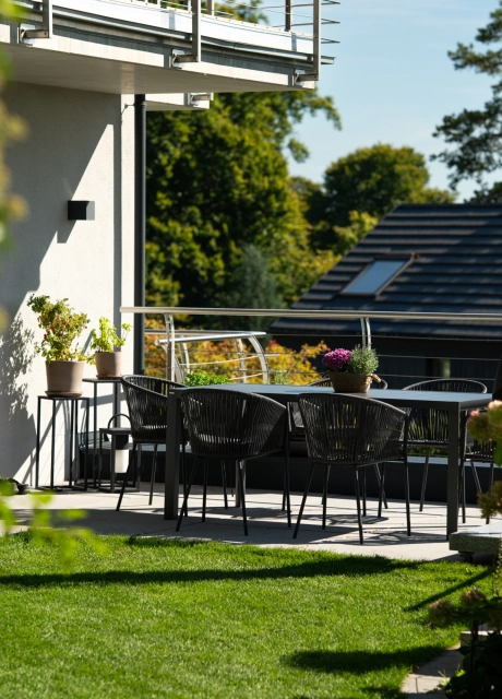 Blick auf eine sonnige Terrasse eines modernen Wohnhauses, auf der ein schwarzer Tisch mit mehreren filigranen Stühlen steht. Die Terrasse ist von dichtem Grün und blühenden Sträuchern umgeben und öffnet sich zu einer gepflegten Rasenfläche. Im Hintergrund sind weitere Gebäude sowie hohe Bäume zu sehen.
