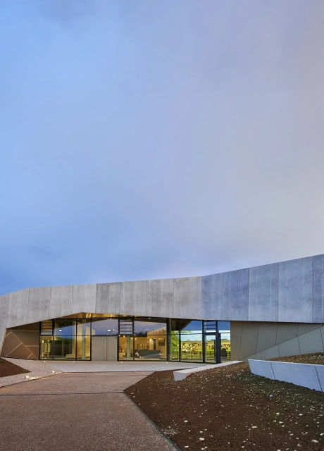 Modernes Gebäude mit Holzoptikfassade und großer Fensterfront. Davor ist ein Weg, welcher auf das Gebäude hinzuführt. Drumherum sind erdige Flächen.