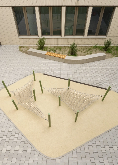 Innenhof eines Schulgebäudes mit großen Fenstern, ausgestattet mit kleinen Klettergeräten und Sitzelementen. Gepflastert ist er mit grau-beigen, versickerungsfähigen Betonsteinen.