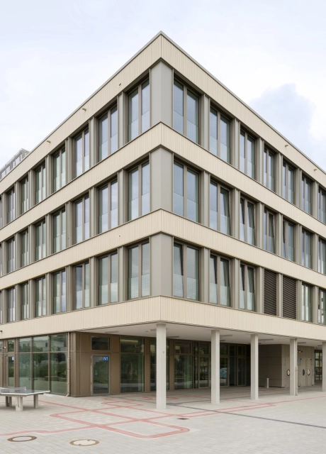 Offener Schulhof mit Tischtennisplatten und einer grau-beigen gepflasterten Fläche. Im Fokus steht ein modernes Schulgebäude mit großen Fenstern.
