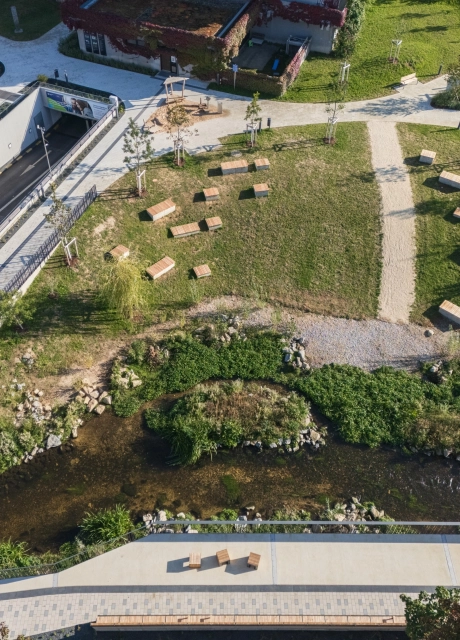 Luftaufnahme eines neu gestalteten Parkareals mit gepflasterten Wegen, einem kleinen Bachlauf, Grünflächen mit vielen Sitzbänken sowie angrenzenden Spiel- und Verkehrsbereichen.