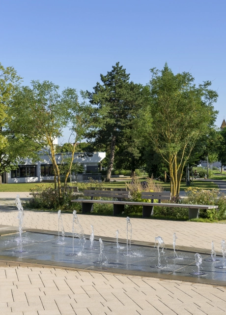 Offener Platz in einer begrünten Park-Umgebung. Inmitten des gepflasterten Platzes ist ein Wasserspiel mit Springbrunnen Elementen platziert. Im Hintergrund sind viele Grünflächen mit Wiesen, Sträuchern und Bäumen. Dahinter ist ein Gebäude mit weißer Fassade zu erkennen.