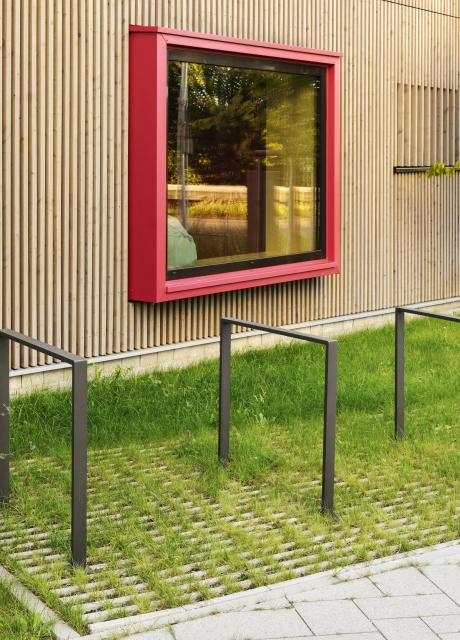 Anthrazitfarbene Fahrradständer auf einer begrünten, mit GDM.SCADA liner, gepflasterten Fläche. Dahinter ist ein Gebäude mit Holzverkleidung und einem großem Fenster mit rotem Rahmen.
