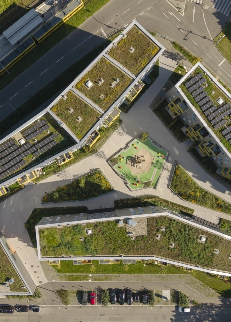 Blick von oben auf große Wohngebäude mit begrüntem Dach. Zwischen den Gebäuden findet sich eine gepflasterte Fläche und grüne Beete. Grünflächen, Parkflächen und Straßen sind außenherum zu finden.