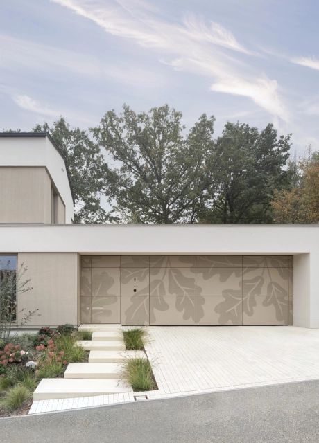 Ein helles, modernen Wohnhaus mit einer gepflegter Einfahrt mit indivdualisierten Garagentoren und einem wild bepflanzten Blumenbeet.