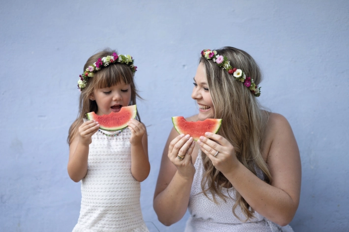 Eine Frau und ein Kind mit Blumenkränzen in den Haaren essen eine Wassermelone vor einer blauen Wand.