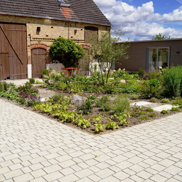 Ein rustikales Backsteingebäude mit großen Holztoren und einen angelegten Garten im Vordergrund. Ein gepflasterter Weg führt durch den Garten, der mit Blumen und Sträuchern bepflanzt ist. Vor dem Garten ist ein naturbeige gepflasterter Hof im Reihenverband angelegt. Rechts ist ein modernes Gebäude mit großen Glastüren zu sehen.
