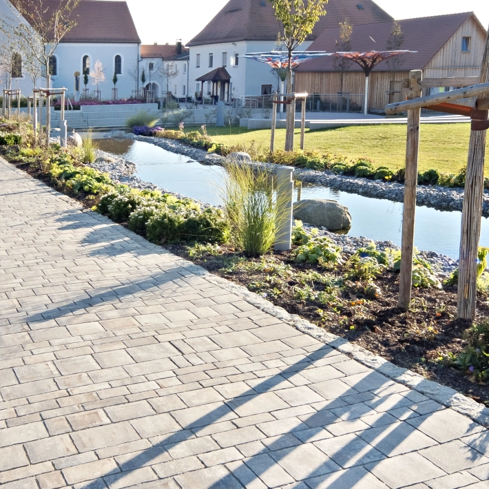 gepflasterter Weg mit neu gestaltetem Dorfplatz in Ursensollen  mit bepflanzen Beeten und angelegtem Bachlauf.
