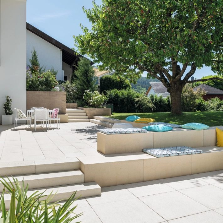 Patio slabs in a landscaped garden with a large tree and concrete seating elements.