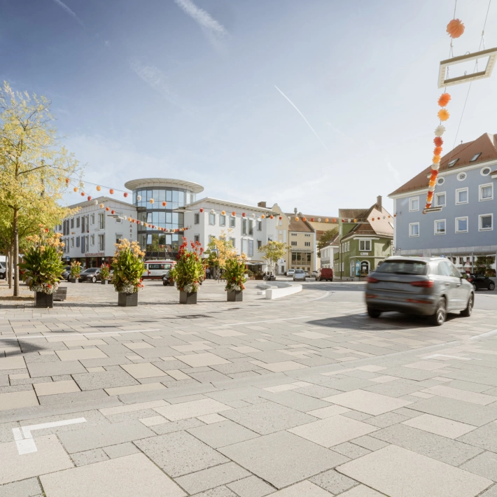 befahrbare Großfromatplatten im wilden Verband und abgestuften Grautönen auf einem Stadtplatz mit Bepflanzung.