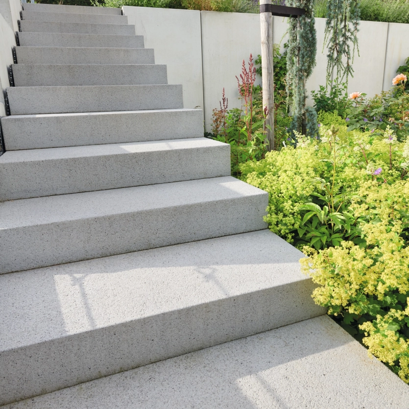 Treppe aus großen, hellen Betonstufen, die zu einer mit bunten Blumen, Gräsern und Sträuchern bepflanzten Gartenfläche führt.