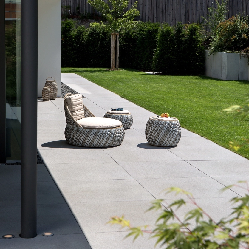 Moderne Terrasse mit grossformatigen, grauen Platten, möbliert mit runden, geflochtenen Sitzmöbeln und einem kleinen Tisch; angrenzend eine gepflegte Rasenfläche und Hecken im Hintergrund.