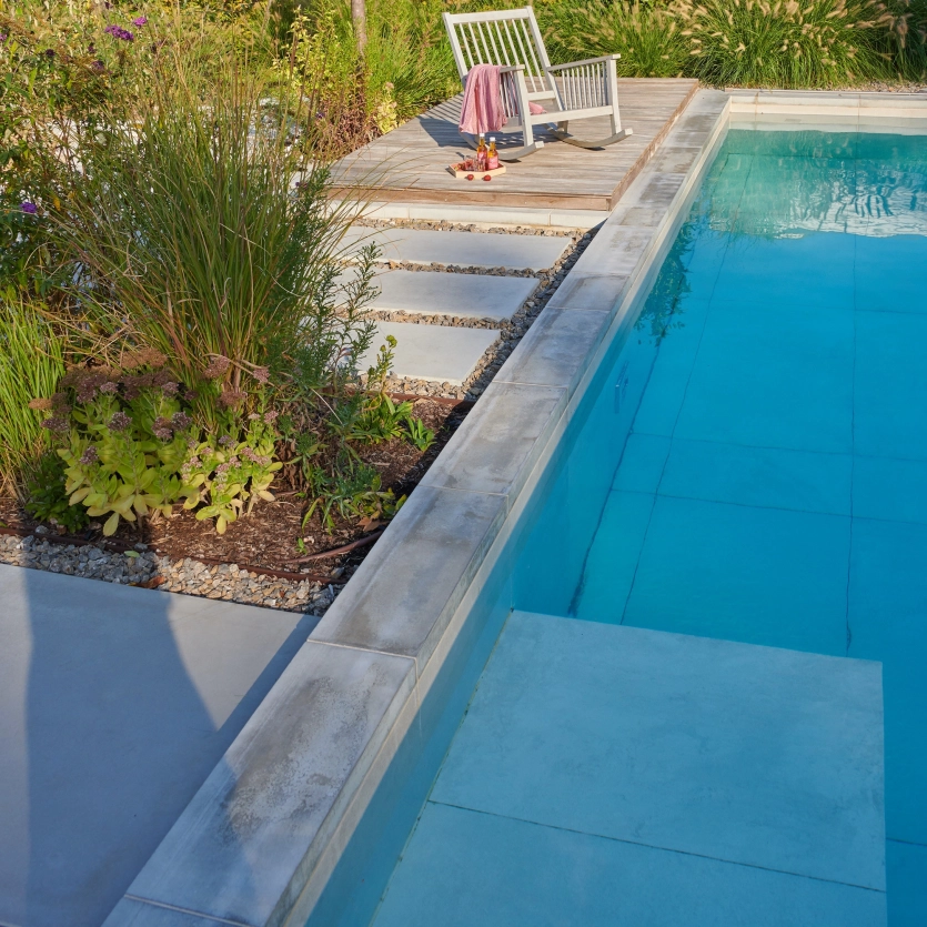 Gemütliche Gartensituation am Pool: Ein Schaukelstuhl lädt zwischen viel Grün ein am Beckenrand den Feierabend zu genießen. Der Terrassenbelag ist mit GDM.MASSIMO ausgestattet.