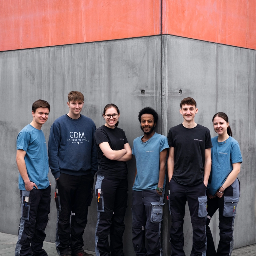 Gruppenfoto der GODELMANN Azubis vor einer grauen Sichtbetonwand, die im oberen bereich einen roten Akzent hat: die sechs Auszubildenden stehen nebeneinander zum Gruppenfoto aufgereiht.