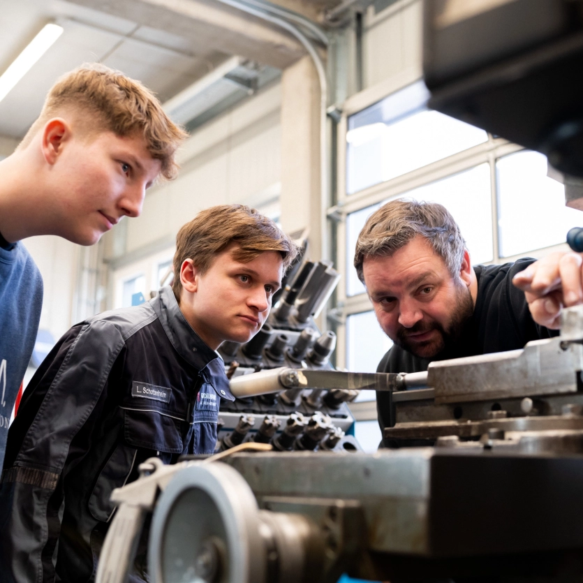 Ausbildungsleiter zeigt zwei Auszubildenden einen technischen Sachverhalt an einer Maschine in der Werkstatt.