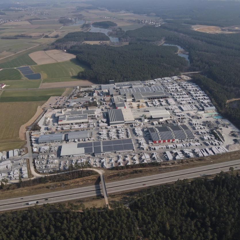 Aerial view of the GODELMANN factory premises in Fensterbach, surrounded by forest areas, fields, and bodies of water. The premises include numerous large buildings, warehouses, and parking lots.