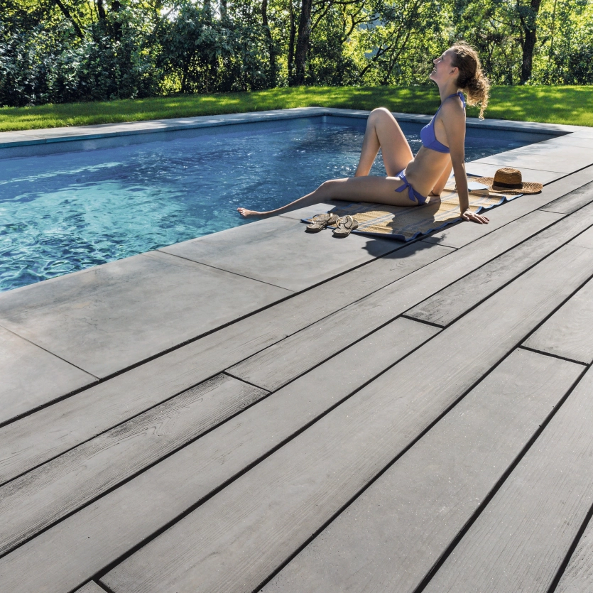 Frau sitzt am Beckenrand und genießt die Sonne. Die Terrasse ist mit Betondielen in Holzoptik von GODELMANN belebt - GDM.MASSIMO plank.