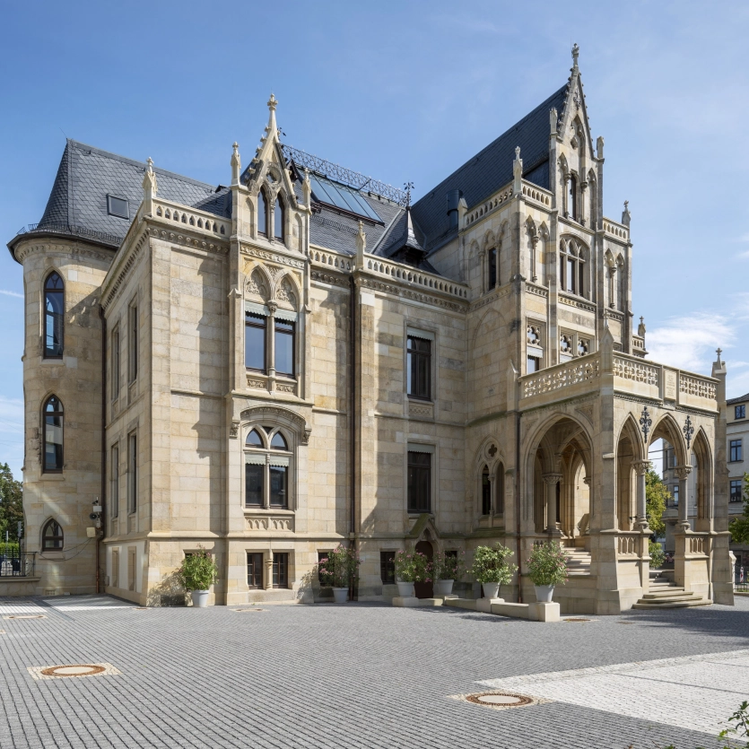 Historische Villa mit mehreren Stockwerken und aufwendigen architektonischen Details. Das Gebäude hat eine helle Sandstein-Fassade, verzierte Fensterrahmen und einen ausgeprägten Eingangsbereich mit darüberliegendem Balkon. Der gepflasterte Platz um die Villa ist gepflegt und von Bäumen und Pflanzgefäßen umgeben. Im Vordergrund sind auch Parkplätze zu erkennen