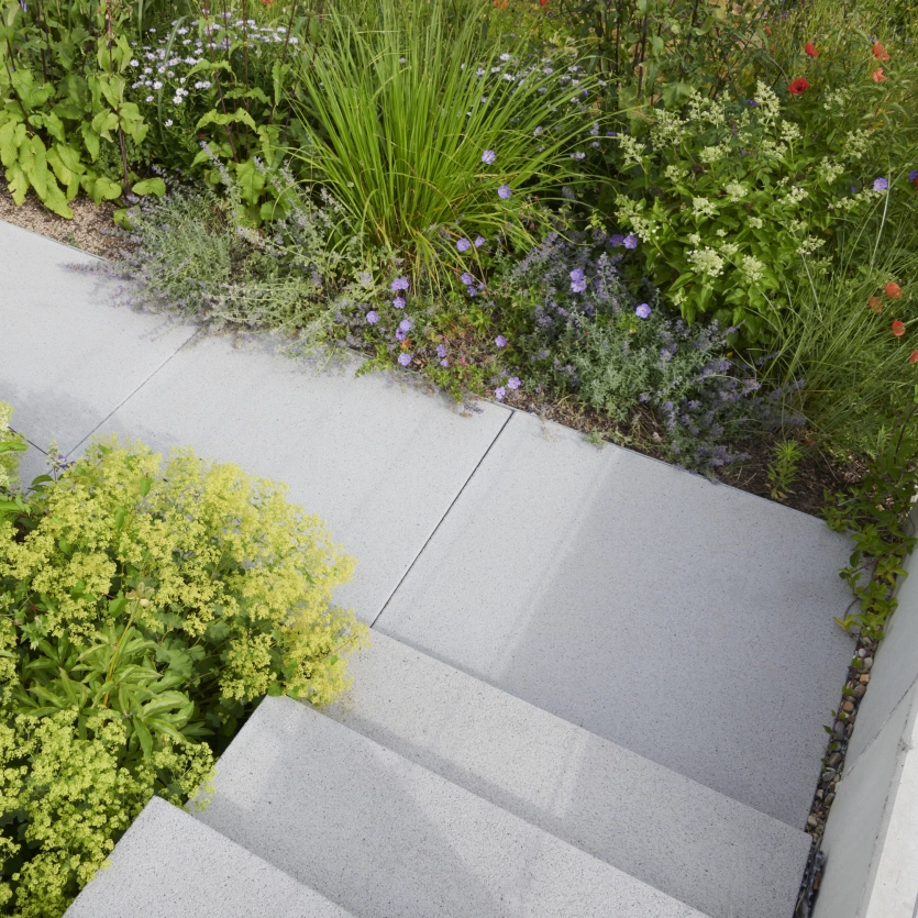 Treppe aus großen, hellen Betonplatten, die zu einer mit bunten Blumen, Gräsern und Sträuchern bepflanzten Gartenfläche führt.
