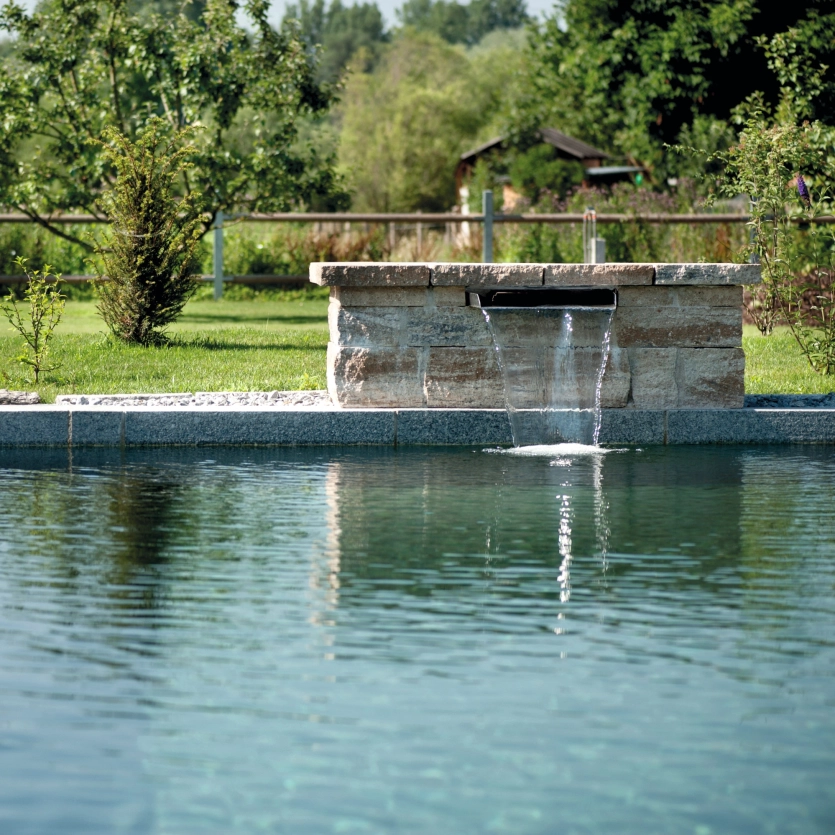 Eine Schwalldusche ist am Rand des Pools in eine GDM.KLASSIK wall-Mauer eingelassen und plätschert ins Wasser.