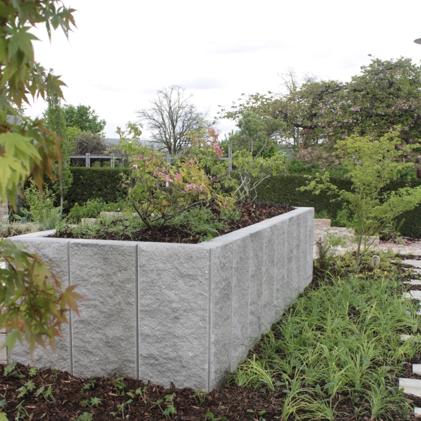 Ein Garten mit einer Stützmauer in L-Form und verschiedenen Pflanzen, die eine grüne und einladende Atmosphäre schaffen.