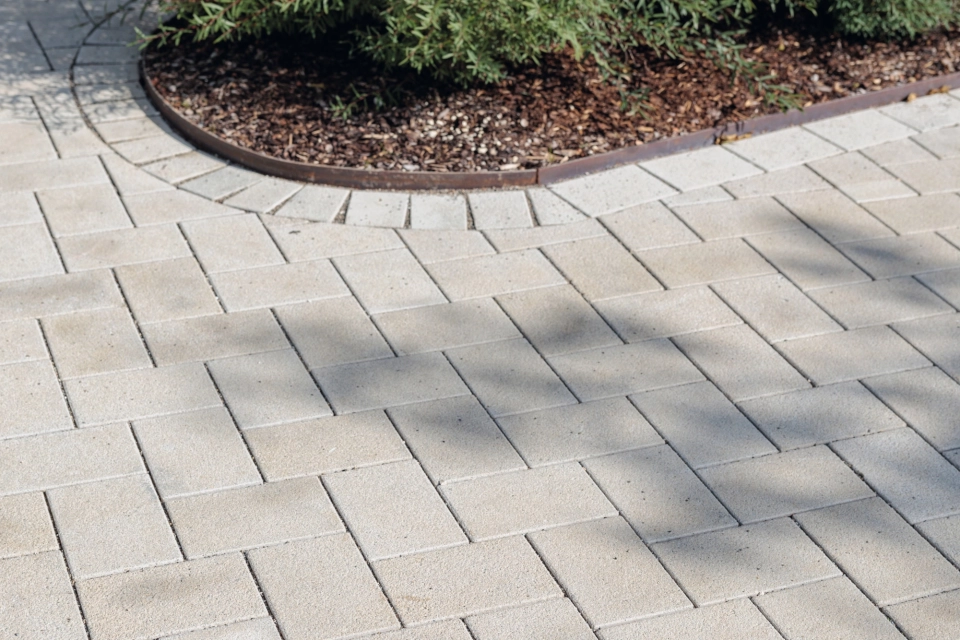 Nahaufnahme eines Weges aus beigem GDM.KLIMASTEIN, der an ein rund angelegtes Beet mit dichtem Grün und dunklem Mulch grenzt. Im oberen Bereich fällt Schatten von umliegenden Elementen auf das Pflaster.