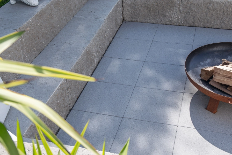 Blick auf eine zweistufige Sitzmöglichkeit aus GDM.MAXI block. In der Mitte der Sitzmöglichkeit steht eine Feuerschale auf grauen GDM.NUEVA light Platten. Im Vordergrund sind die hellgrünen Blätter einer Pflanze.