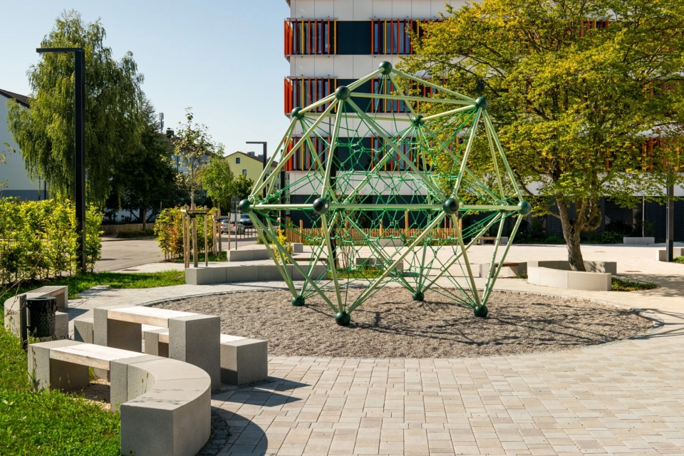 Kalk-Grau und Beige gepflasterter Schulhof mit vielen Sitzelementen und Grünflächen an einem sonnigen Tag mit blauem Himmel. Ein Klettergerüst innerhalb einer Sandfläche ziehrt den Hof und große Bäume spenden Schatten. Im Hintergrund steht ein Schulgebäude mit weißer Fassade und orange-gelben, geometrischen Verzierungen