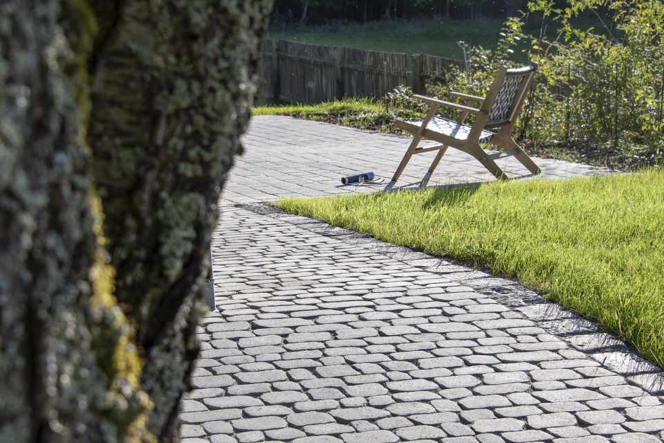 Im Vordergrund verläuft ein hell gepflasterter Weg aus Betonstein, der an einer Sitzbank vorbeiführt und von Rasen sowie alten Bäumen eingerahmt wird, während ein Holzzaun im Hintergrund die ruhige Gartenlandschaft abschließt.