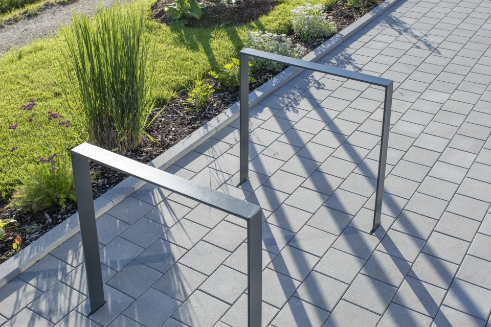 Nahaufnahme von anthrazitfarbenen Fahrradständern auf grauem Betonsteinpflaster. Im Hintergrund ist eine hügelige Landschaft mit Sträuchern, Gräsern und Bäumen zu erkennen.