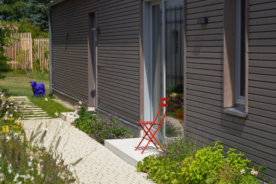 A modern building with a façade of dark brown wooden panels. A red folding chair adorns the pedestal of the entrance door and a natural beige paved path in a row separates the building from a garden in the foreground. A few conifers can be seen in the background. 