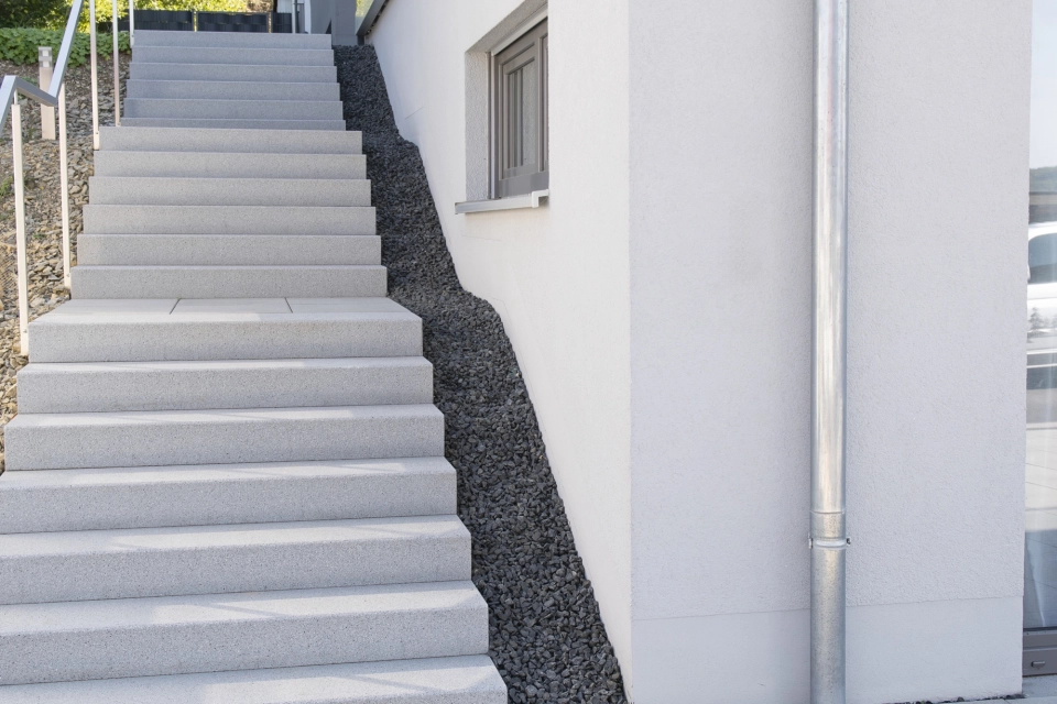 Blick auf eine helle Treppe mit Blockstufen, welche an einer hellen Gebäudefassade entlang führt. Davor ist eine mit GDM.TETRAGO stone gepflasterte Fläche.